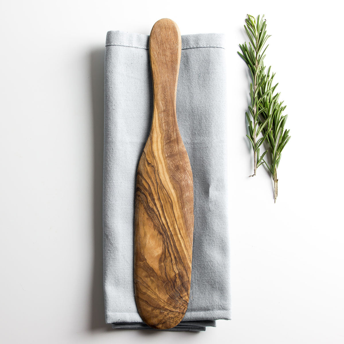 olive wood spatula resting on a grey napkin on a plain white surface with some herbs alongside