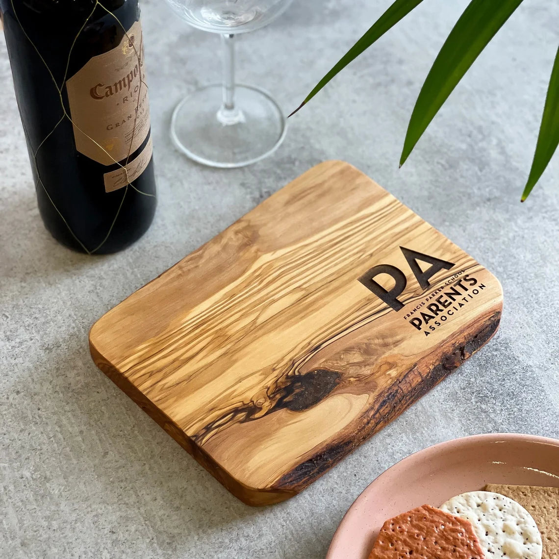 An olive wood chopping/cheese board with a branded logo engraved on it, displayed on a table with some food items.