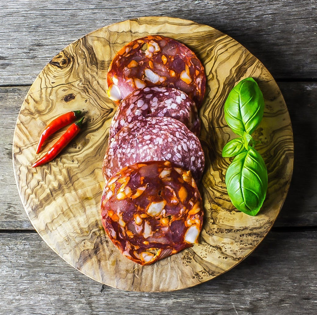 a round olive wood board with some chillies, salami and basil resting on the top on a grey surface.