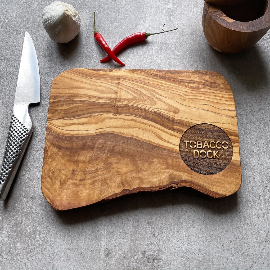 An olive wood chopping/cheese board with a branded logo engraved on it, displayed on a table with some food items.