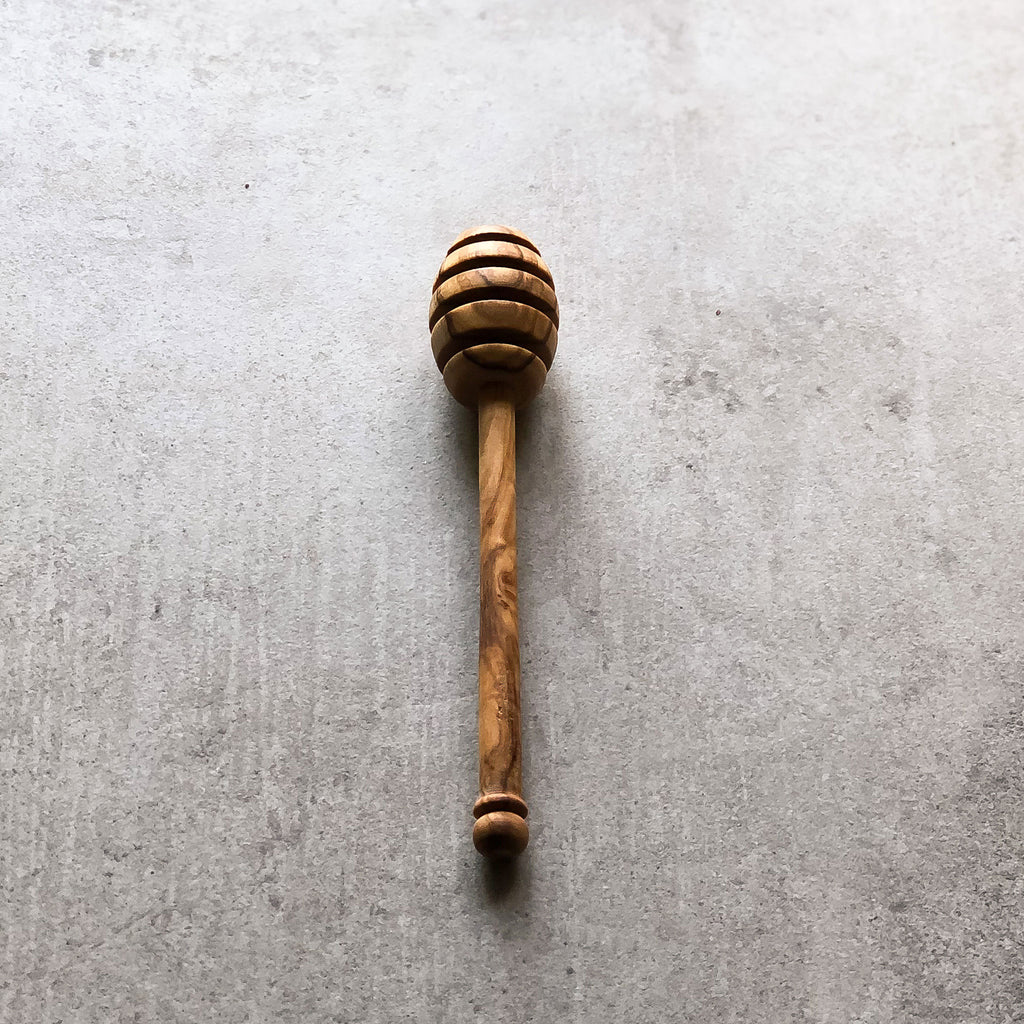 A honey drizzler made of olive wood, with a handle, placed on a grey surface.