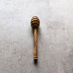 A honey drizzler made of olive wood, with a handle, placed on a grey surface.