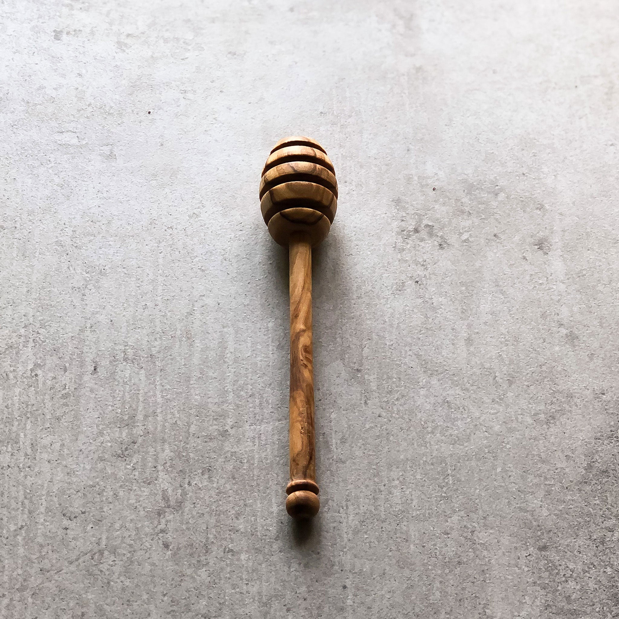 A honey drizzler made of olive wood, with a handle, placed on a grey surface.