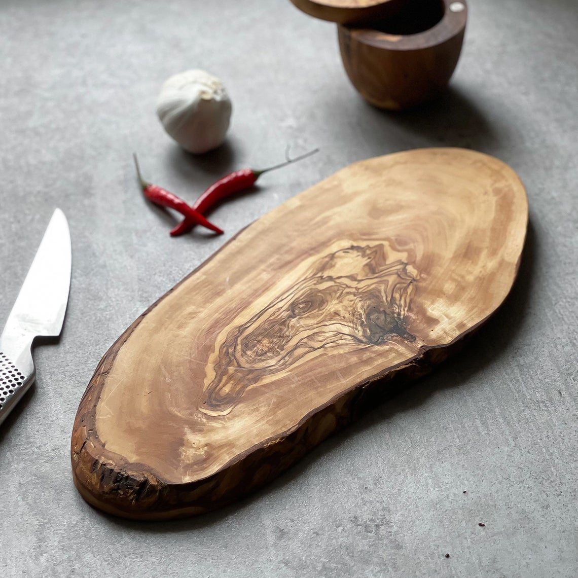 a rustic olive wood board on a grey surface with some chillies, garlic, a knife and salt pot