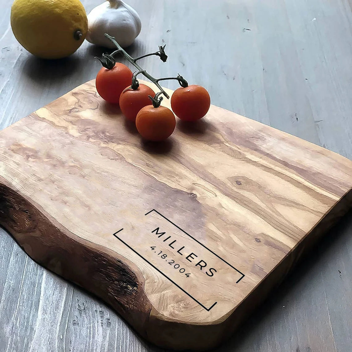A rustic olive wood cutting board with a personalized engraving, accompanied by tomatoes, lemon, and garlic on top, set on a wooden table.