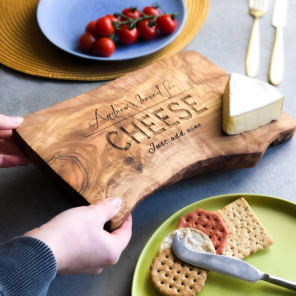 Wooden cutting board with engraved text being placed with cheese on and surrounded by crackers and cherry tomatoes. The surface is grey and also features some gold cutlery.